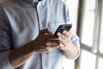 Hand of young business man in casual using virtual app, online service on smartphone, texting, chatting online, typing message, touching screen, standing at window, holding mobile phone. Close up