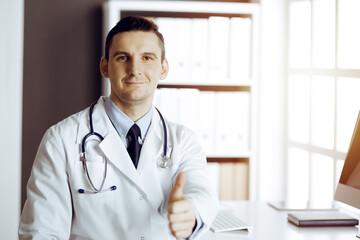 Friendly male doctor sitting and showing OK sign with thumbs up at his working place in sunny clinic. Perfect medical service in hospital - 459277988