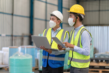 Asian male supervisor engineer in helmet safety wearing hygienic mask to protect coronavirus hold laptop talking with technician working at warehouse factory industrial.