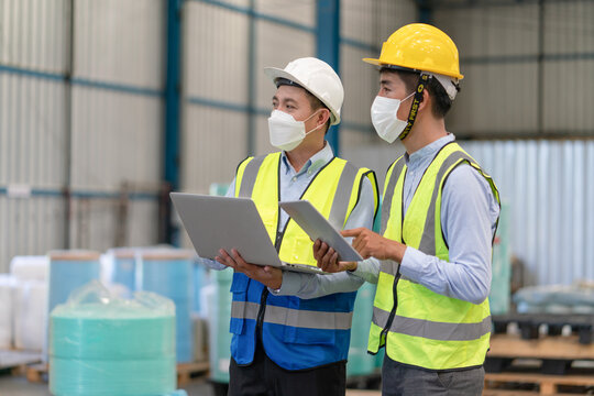 Asian Male Supervisor Engineer In Helmet Safety Wearing Hygienic Mask To Protect Coronavirus Hold Laptop Talking With Technician Working At Warehouse Factory Industrial.