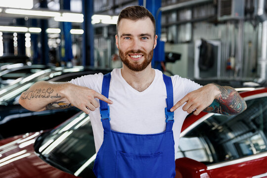 Strong smiling young male professional technician car mechanic man in denim blue overalls white t-shirt point on himself work in light modern vehicle repair shop workshop indoors Tattoo traslate fun.