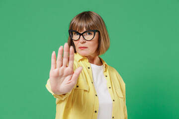 Elderly strict sad serious caucasian woman 50s in glasses yellow shirt showing stop gesture with...