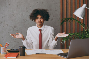 Young mistaken confused uncertain african american employee business man in classic shirt tie spread hands like oops gesture sit work at white office desk with pc laptop Achievement career concept