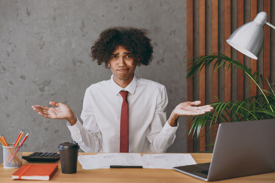 Young mistaken confused uncertain african american employee business man in classic shirt tie spread hands like oops gesture sit work at white office desk with pc laptop Achievement career concept