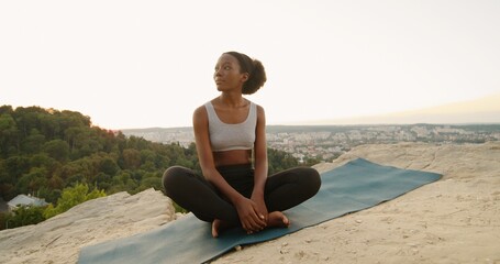 Fototapeta premium Young multiracial woman sitting in yoga posture and meditating at the mountains. Girl performing aerobics exercise and morning meditation at the nature. Physical and spiritual practice concept