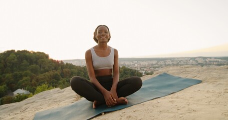 Fototapeta na wymiar Young multiracial woman sitting in yoga posture and meditating at the mountains. Girl performing aerobics exercise and morning meditation at the nature. Physical and spiritual practice concept