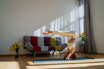 Young female in white sportswear practicing yoga headstand exercise indoors