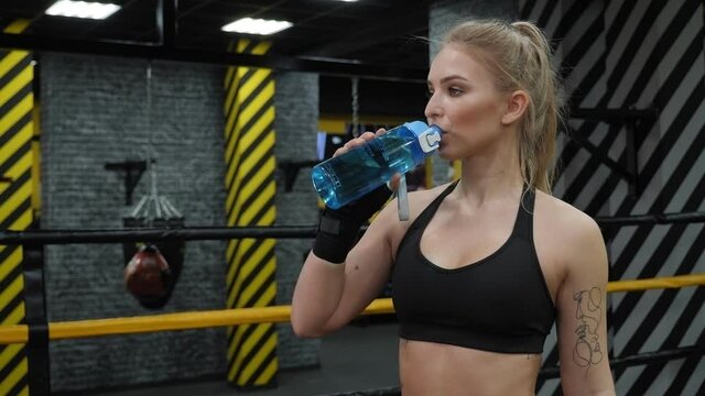 A tired girl drinks water while standing in a boxing ring during a boxing training session.