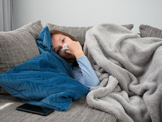 A sick young, cold woman wrapped in a soft blanket is lying on the couch at home. The girl with a runny nose is blowing her nose into a handkerchief.