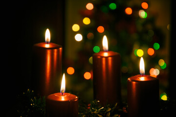 Four red candles burning on advent wreath with blurred colorful christmas tree with glowing lights in the background on evening. Merry Christmas, Advent crown decoration, 4th sunday, Xmas backdrop