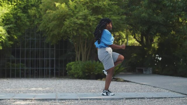 Tracking shot of chubby woman doing high knees running exercise. Long shot of Afro-American female person in sportswear doing fat burning outdoor workout. Cardio, sport, body positive concept