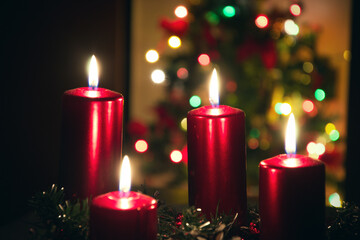 Four red candles burning on advent wreath with blurred colorful christmas tree with glowing lights in the background on evening. Merry Christmas, Advent crown decoration, 4th sunday, Xmas backdrop