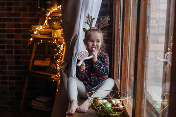 Little beautiful girl in a Christmas headdress sits by the window and eats marshmallows. Christmas concept. Smiling funny girl in deer antlers in the studio.