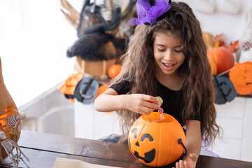 cute little girl in costume of witch holding pumpkin jack with candies, having fun in kitchen, celebrating Halloween