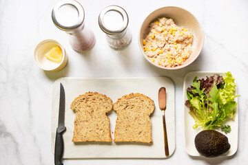 Preparing healthy sandwiches; super grains sliced bread, tuna, corn, avocado, lemon and vegetables on marble cutting board. Top view
