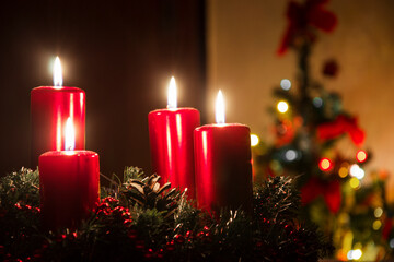 Four red candles burning on advent wreath with blurred colorful christmas tree with glowing lights in the background on evening. Merry Christmas, Advent crown decoration, 4th sunday, Xmas backdrop