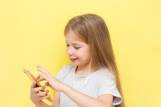 A Funny Little Girl With Long Hair On A Yellow Background Holds A Smartphone In Her Hands With Headphones And Smiles. The Concept Of Children And Gadgets, Social Networks, Tik Tok.