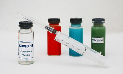 A group of multicolored glass jars with the Covid-19 coronavirus vaccine and a syringe on a white surface.