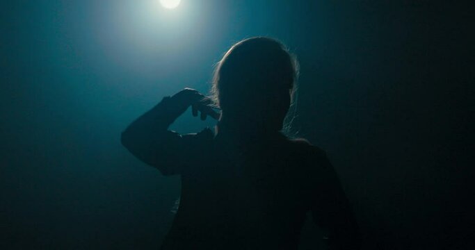 Beautiful experienced talented woman with long blonde hair dances on a dark stage lit by a lamp, practicing new moves, poses, moving sexily, the shadow of the girl's silhouette