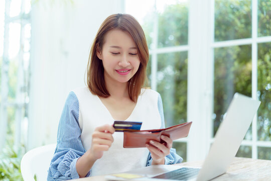 A beautiful Asian woman wearing a white shirt with a credit card comes out of her wallet smiling happily in front of her laptop preparing for online shopping.