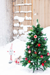 Artificial fir tree decorated with Christmas red toys outdoors in yard in snowy background with dwarf, bag with gifts and falling snow.  New Year and Christmas concept