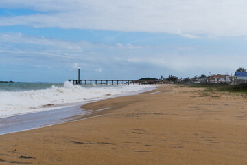 View of the beach of Rio das Ostras in Rio de Janeiro with the Pier of Emissary. Sunny day, blue sky and some clouds. Strong sea and yellowish sand and lots of rocks.