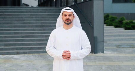 Portrait view of the bearded arabian man wearing kandura looking at the camera and smiling with pleasure while spending time at the street near his office