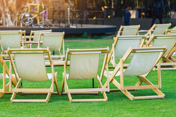 Many empty white deck chairs with tables in lawn is surrounded by shady green grass. Comfortable on outdoor patio chairs in garden.  Lawn chairs in the park. Sunbeds in the garden. Selective focus.