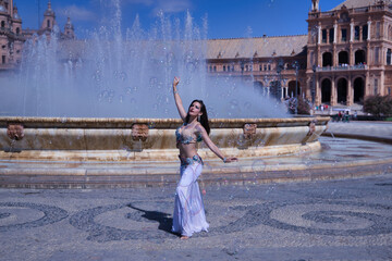 Obraz premium Middle-aged Hispanic woman, wearing a beautifully colored dress with rhinestones, for belly dancing, dancing surrounded by soap bubbles in front of a water fountain. Belly dance concept.