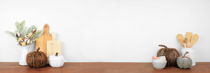 Fall theme kitchenware, rustic pumpkin decor and utensils on a wooden shelf or counter. Banner against a white wall background with copy space. Autumn kitchen home cooking concept.