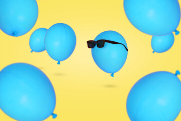 Blue balloon in sunglasses floating in the air with other balloons against blue background.