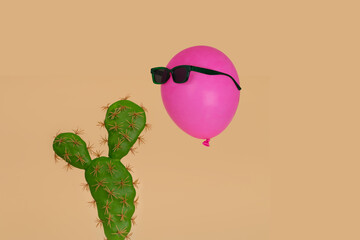 Cactus plant with above it floating a pink balloon in sunglasses on a beige background.