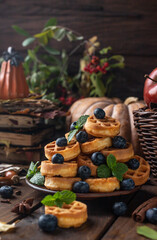 many waffles are stacked on a plate, decorated with mint leaves and blueberries. Vegetables are lying around on a wooden table
