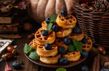 many waffles are stacked on a plate, decorated with mint leaves and blueberries. Vegetables are lying around on a wooden table