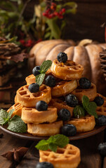 many waffles are stacked on a plate, decorated with mint leaves and blueberries. Vegetables are lying around on a wooden table