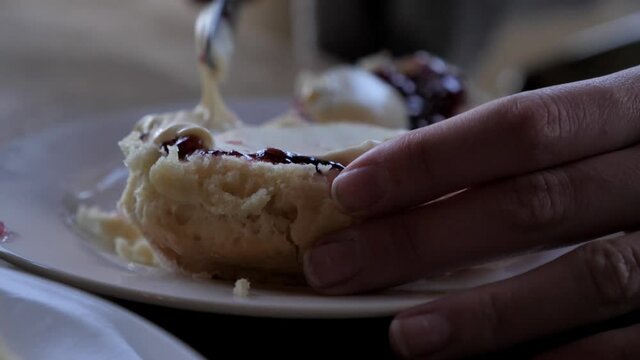 Slow plan around female hand spreading thick cream onto jam covered traditional bakes scone with silver metal knife on white plate for afternoon tea.