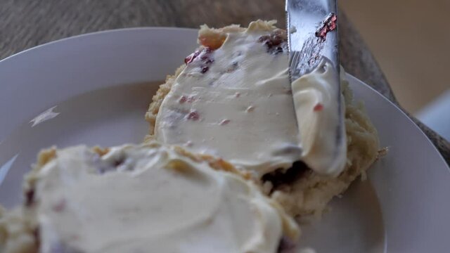 Person spreading whipped thick cream over homemade traditional baked jam scone with silver metal nice on white plate and timber table in rustic setting for afternoon tea.