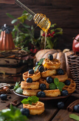 many waffles are stacked on a plate, decorated with mint leaves and blueberries. On top of them, honey pours from glass spoons