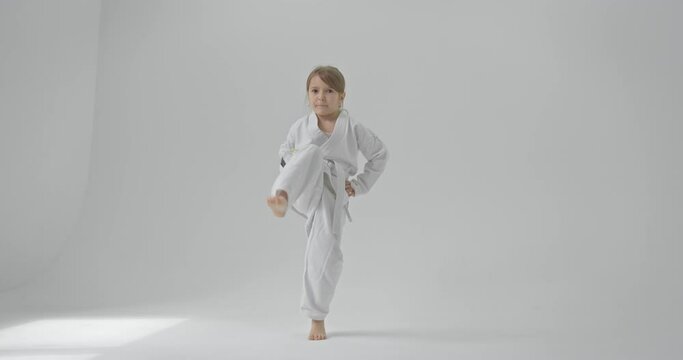 Concentrated child looks at the camera, kicks and kicks her to the ground. Baby in a white kimono on a white background.