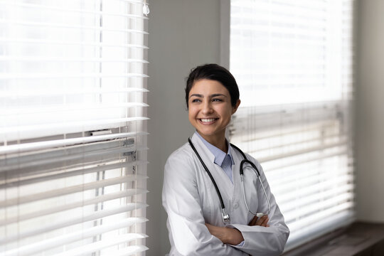 Confident female Indian doctor standing by window, looking away with happy smile. Young general practitioner, therapist thinking of future career vision, enjoying success. Head shot portrait