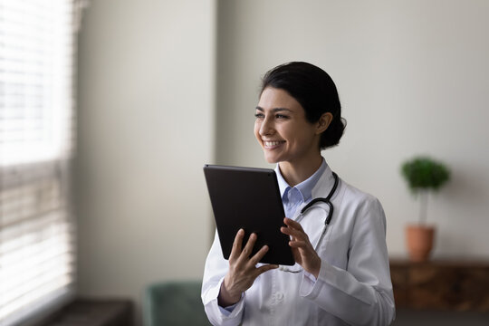 Happy young female Indian doctor holding tablet computer, looking at window away, smiling, thinking. General practitioner, therapist using gadget for online consultation. Head shot portrait