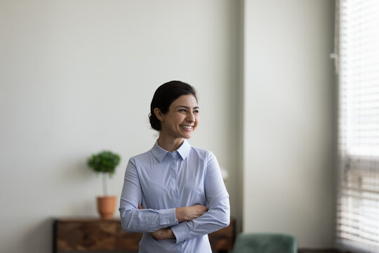 Happy millennial Indian businesswoman looking at window with folded arms, smiling at good thought, thinking of business project future vision. Ambitious employee dreaming of career. Head shot portrait