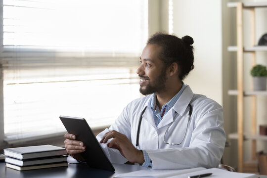 Happy millennial Afro American doctor looking at window away, smiling, holding tablet, thinking over distance online consultation. Practitioner using gadget for reading electronic medical records