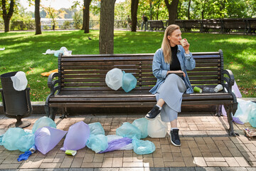 Woman sitting at the empty bench with a lot of plastic bags on it and drinking coffee