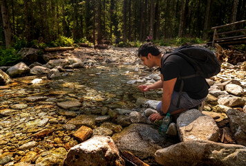 Brown Indian arab man with backpack sitting next to clean water stream or river drinking water from it, taking break while trekking or hike at forest creek . Adventure in nature during summer day.