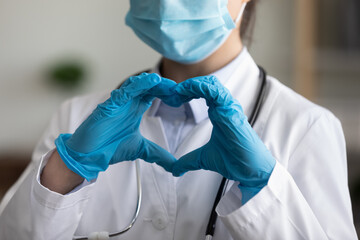 Female doctor in face mask, protective latex gloves showing finger heart shape at camera. practitioner expressing love, support to medical coworkers, covid patients. Pandemic, healthcare. Close up