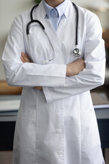 Cropped view of female general practitioner wearing white coat, standing and posing with folded hands. GP doctor, therapist, physician, medical specialist, medic employee in uniform close up shot