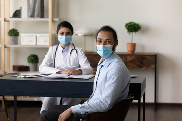 Portrait of happy multiethnic female doctor and male patient wearing face masks at appointment, sitting at practitioner table, looking at camera both, smiling. Medic care on epidemic, healthcare