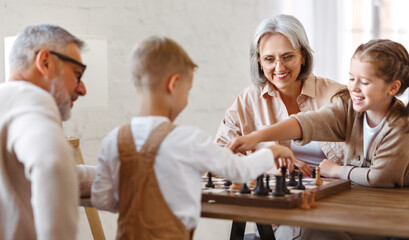 Fototapeta premium Children brother and sister playing chess while sitting in living room with senior grandparents