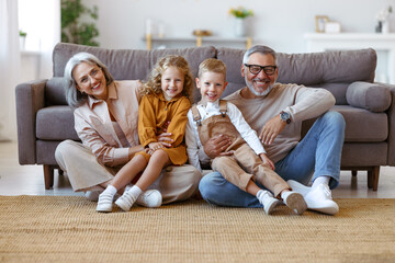Happy family grandparents and two little kids embracing and smiling at camera while sitting on floor